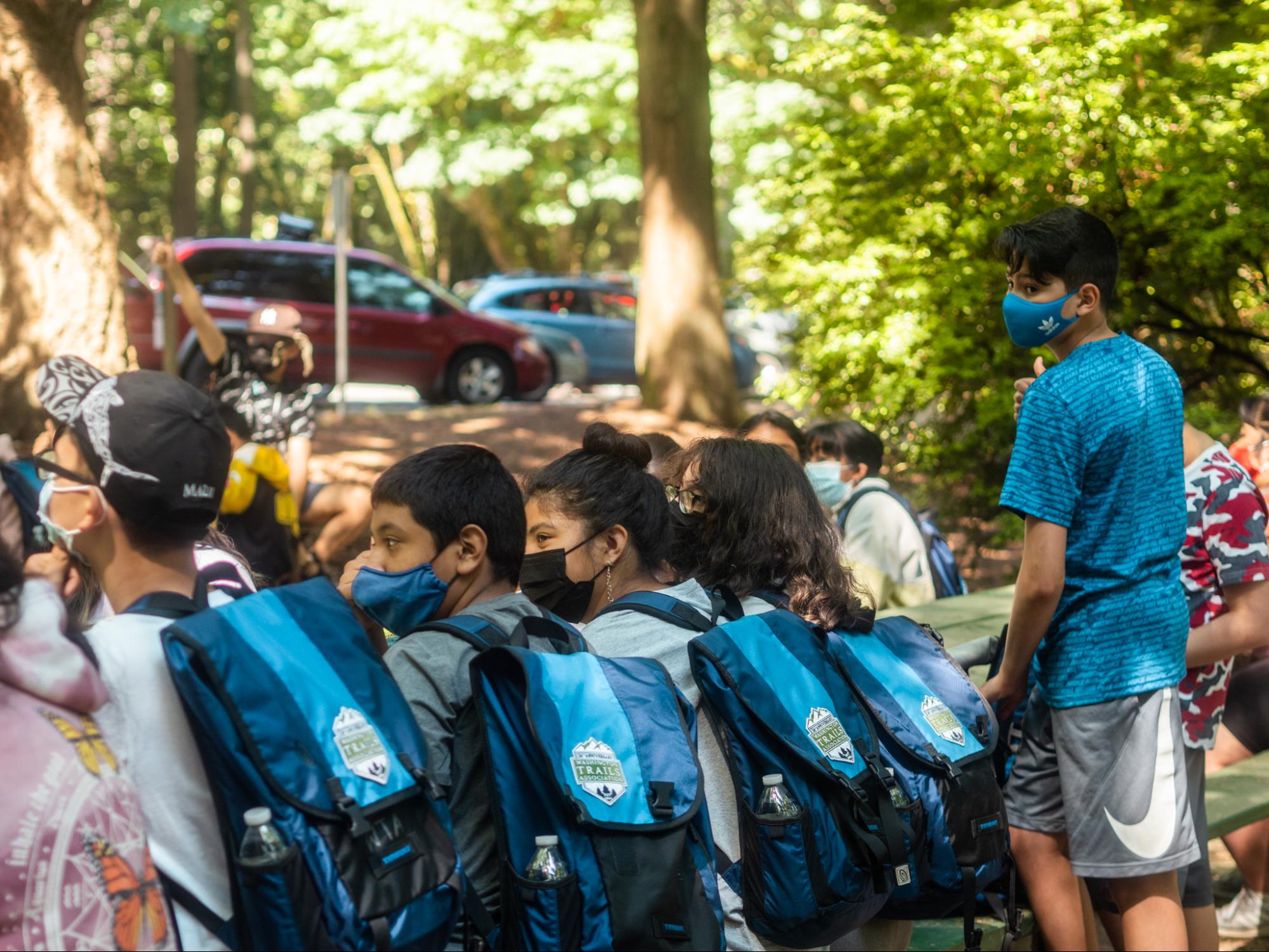 El Centro De La Raza A group of kids wearing masks and blue WTA backpacks wait to head out on a hike.