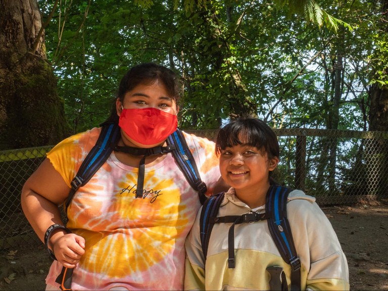 El Centro de la Raza Two students, both wearing backpacks, stand in front of trees and a small fence and smile at the camera.