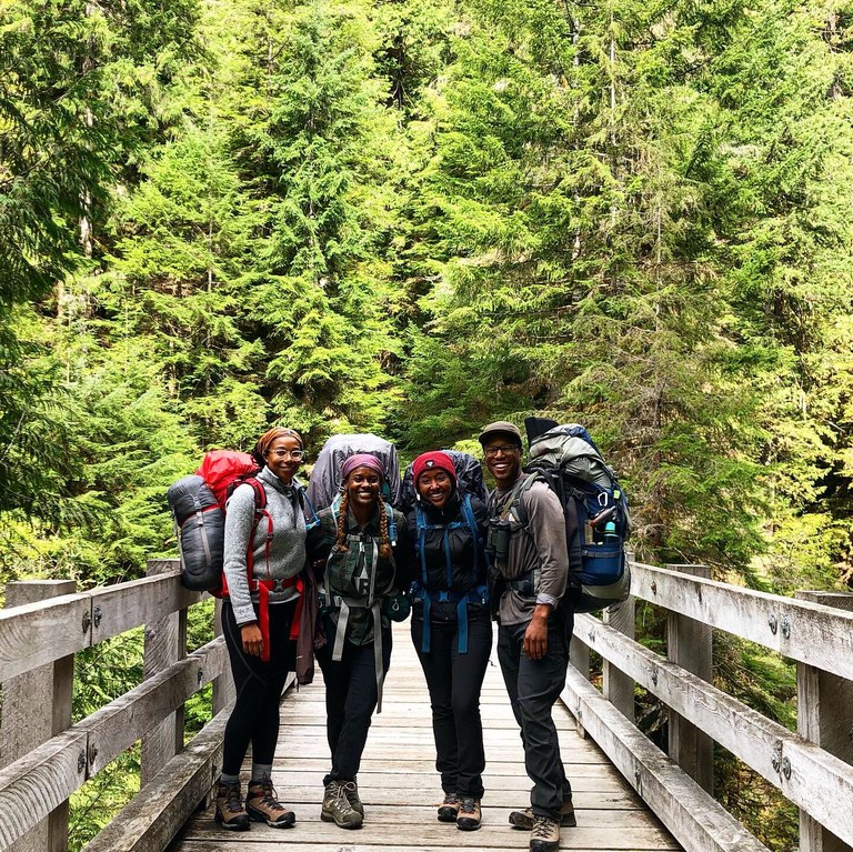 Four hikers standing together on a bridge and smiling. All are wearing backpacking packs. 