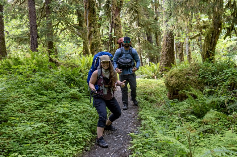 Two hikers with huge smiles and big backpacks pose for a photo on a trail winding through a lush forest. 