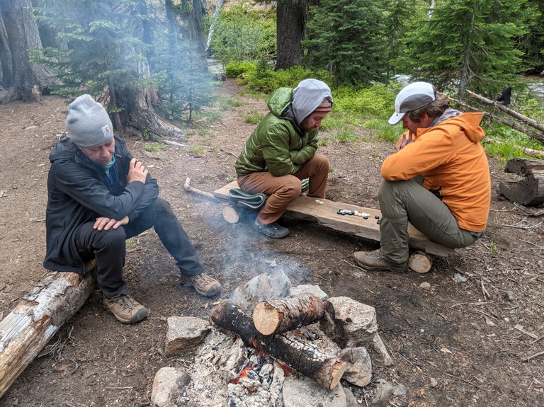 Entiat River BCRT. Photo by Ryan Woods. Two crew members on a backcountry response team trail work party on the Entiat River Trail playing Hive board game on a log with another crew member watching the campfire. Photo by Ryan Woods.