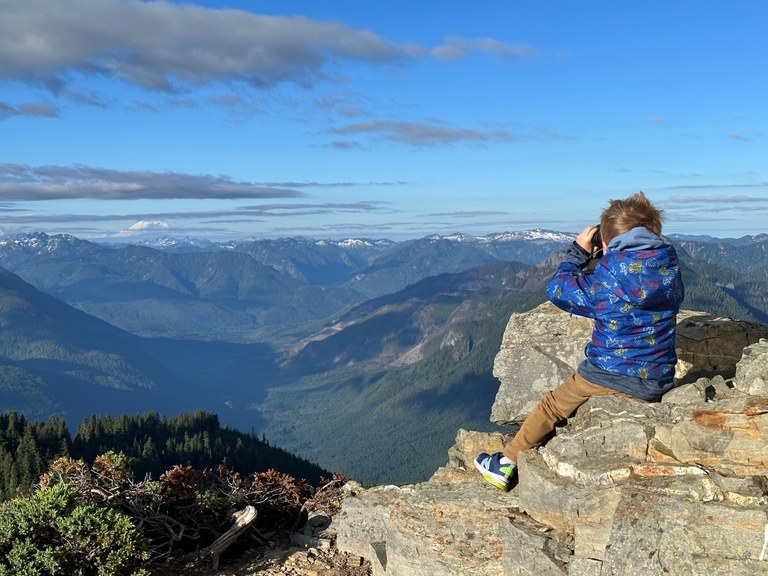 A child sits at the summit of Evergreen Mountain Lookout looking at Mount Rainier through binoculars. Photo by barnacles.