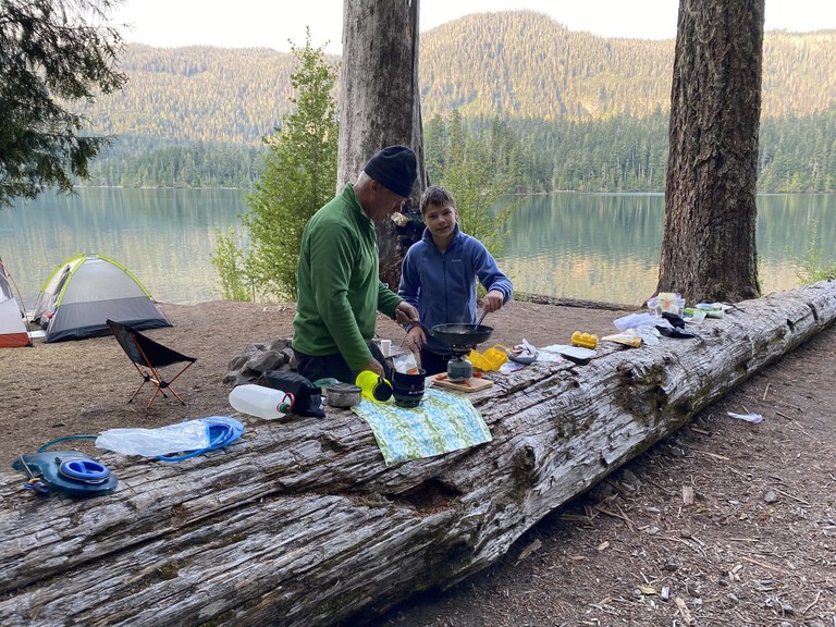 A teen cooks over a backpacking stove, with his grandfather standing nearby. The stove and other kitchen equipment are spread out along a large fallen log. Two tents and a lake are in the background. 