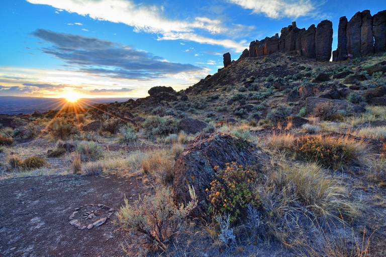 Columns of dark basalt rise in the back of the photo with sagebrush in the front as the sun sets. 