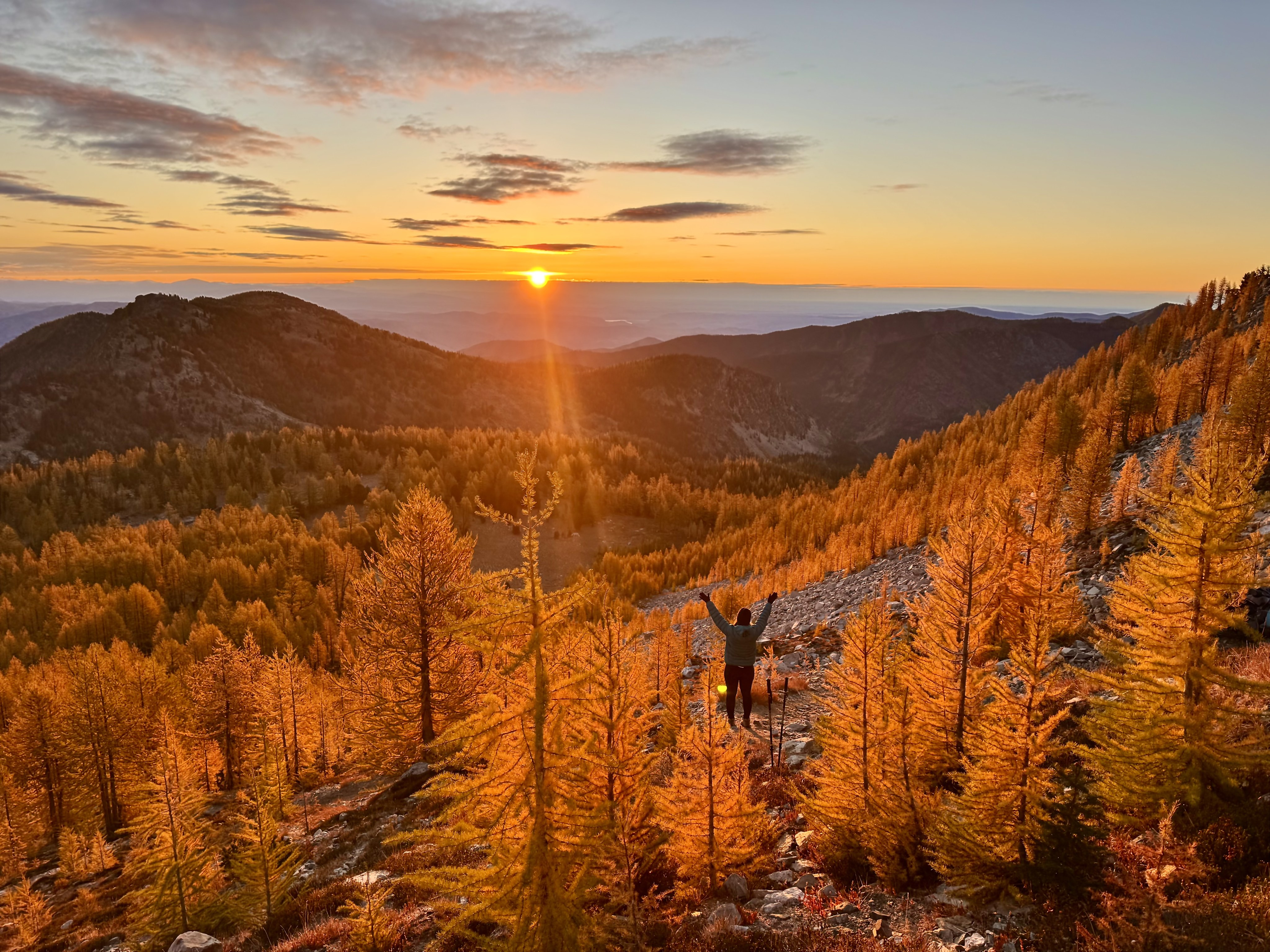 The sun pops over a ridge where a hiker stands with arms outstreched among glowing larches