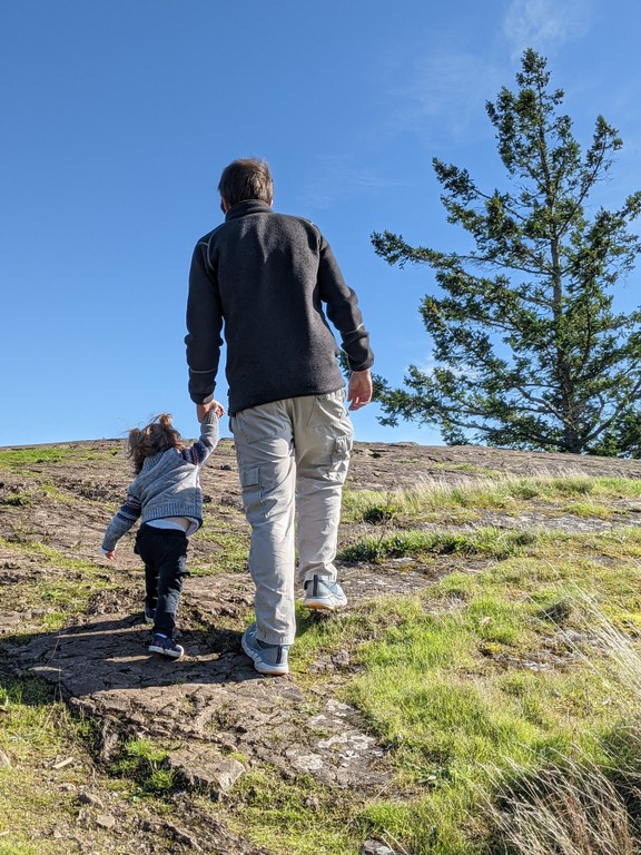 A hiker walks up a steep rock, holding the hand of a toddler, with blue sky and an evergreen tree in the background.
