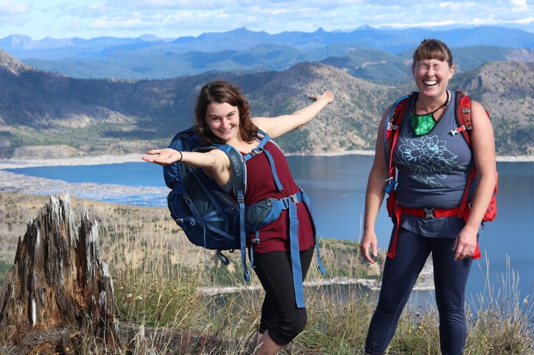A mother and her adult daughter pose for a photo together. The daughter has her arms wide and is smiling at the camera. Her mother is laughing with her eyes closed and a huge smile. A lake with a raft of floating is visible in the background.