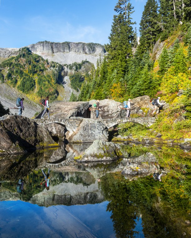WTA volunteers walk over an arching stone bridge that crosses over a subalpine lake.