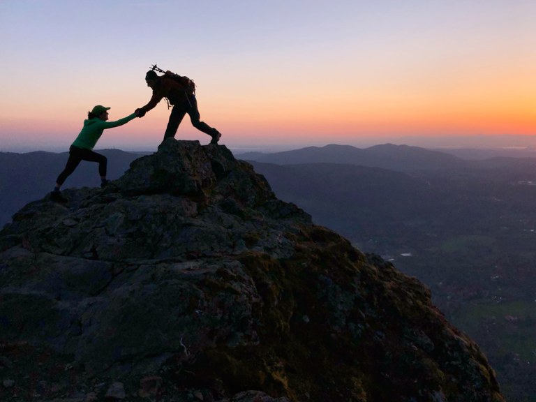A hiker holds out there hand to help another hiker step up onto a rock promontory. 