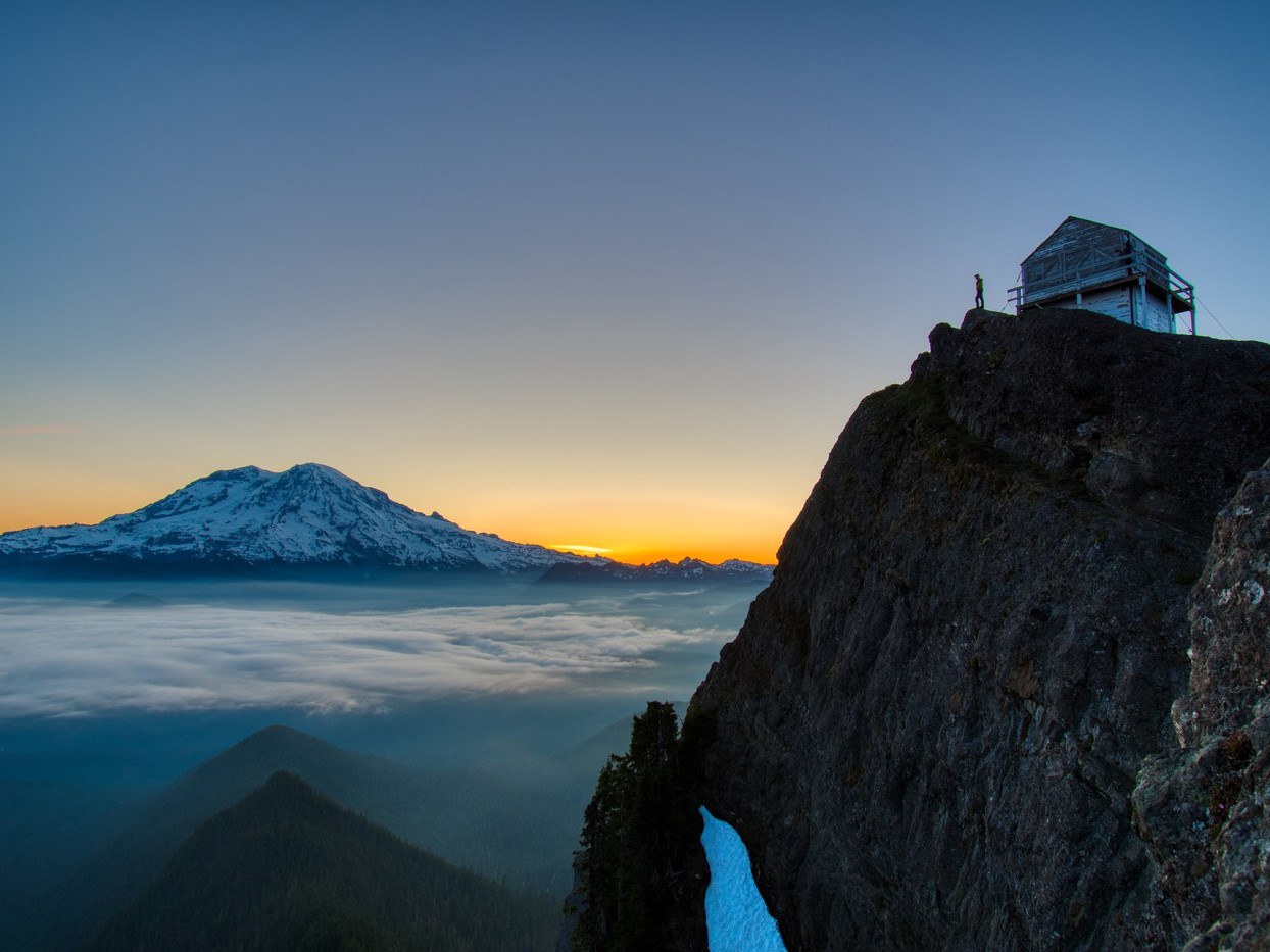 Hiker at a fire lookout looks over cloudy valley and an orange sky behind Mount Rainier
