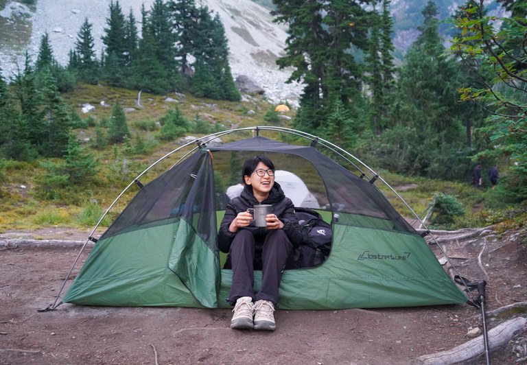 Person smiling holding cup inside of green tent with forest in background.