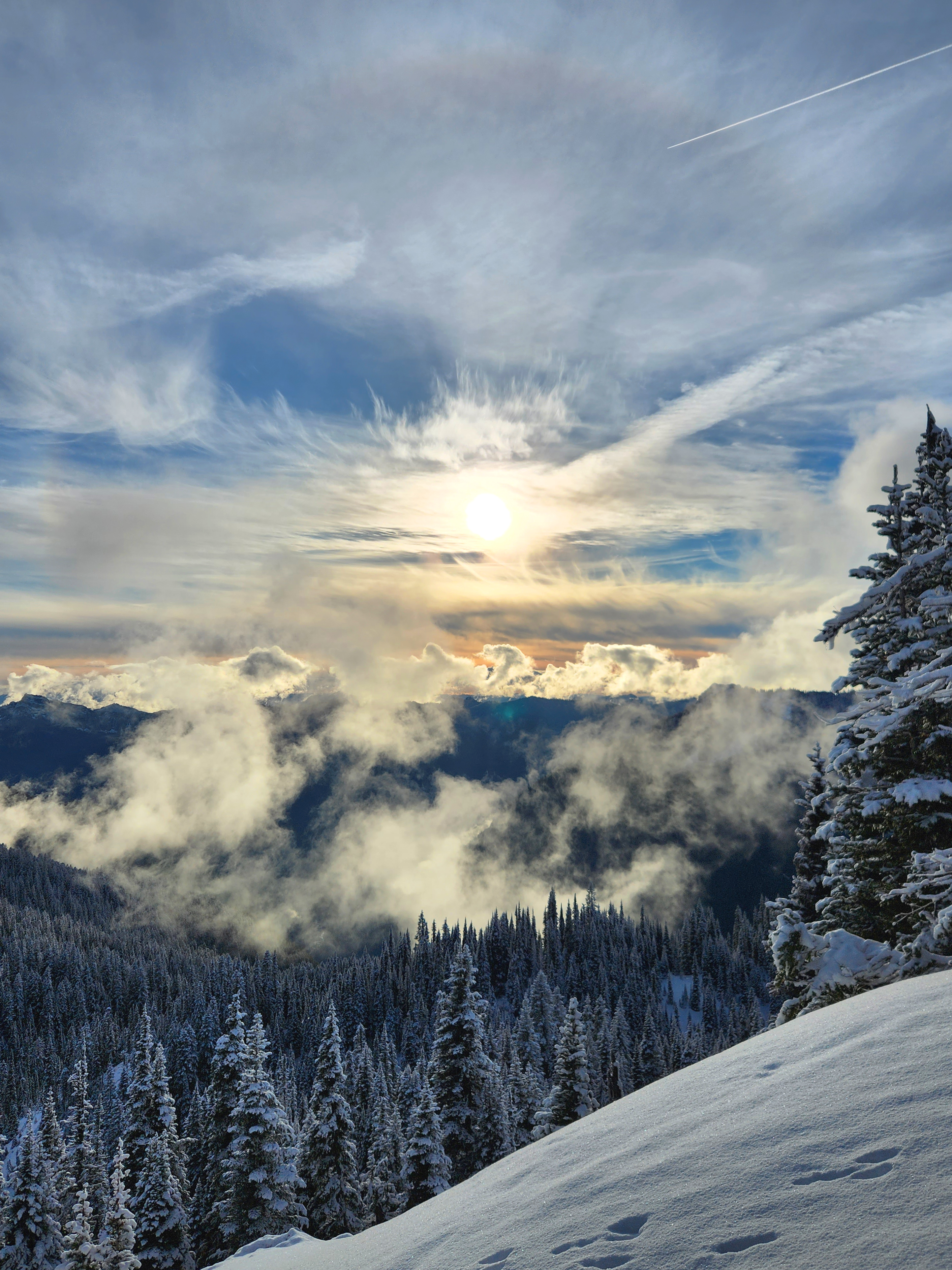 Looking over a snowy hill top, blue sky, wispy clouds,  snow covered trees and mountains.