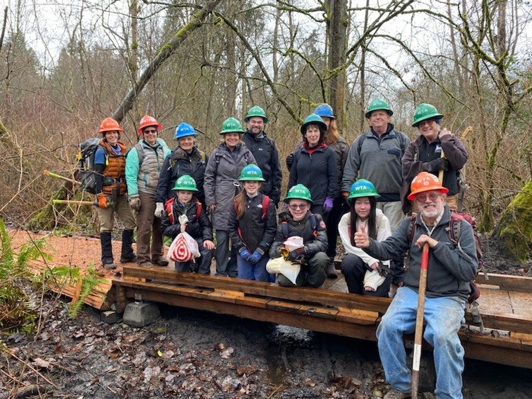 A volunteer trail crew stands on the new bridge they just built. The crew consists of green hats, blue hats, and a few orange hats.
