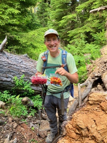Mike stands on trail in front of some greenery holding some fresh veg and smiling. 