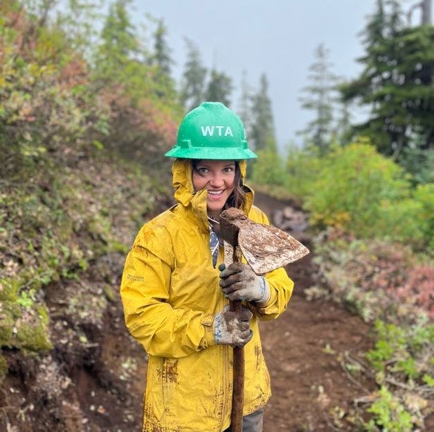 A trail maintainer in a rain jacket holds a tool and smiles.