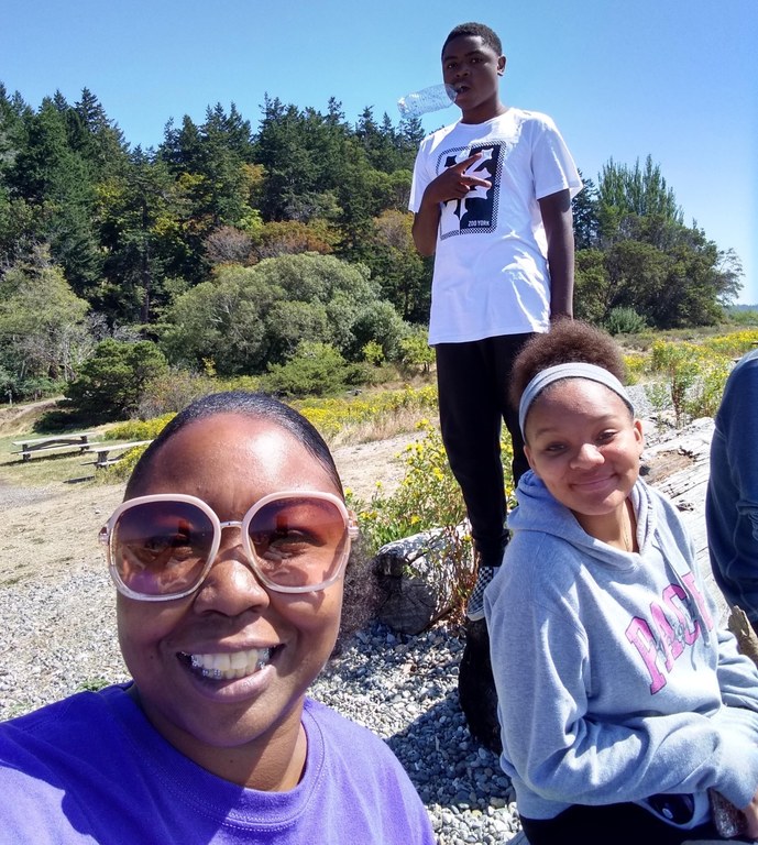Renee Pierce A woman with sunglasses with white frames and a huge smile takes a selfie with two teens. Green trees and blue skies are in the background.