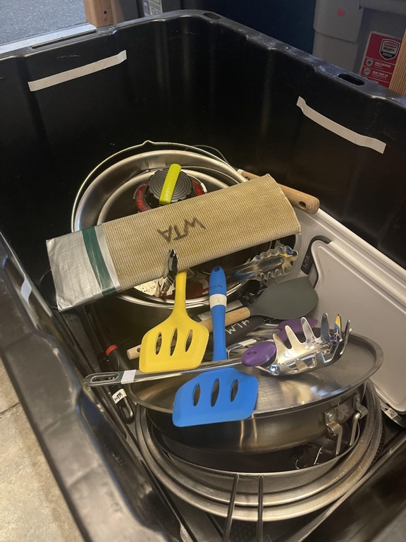 A black box containing cooking supplies for camping trips including several utensils which are haphazardly scattered through out the box.