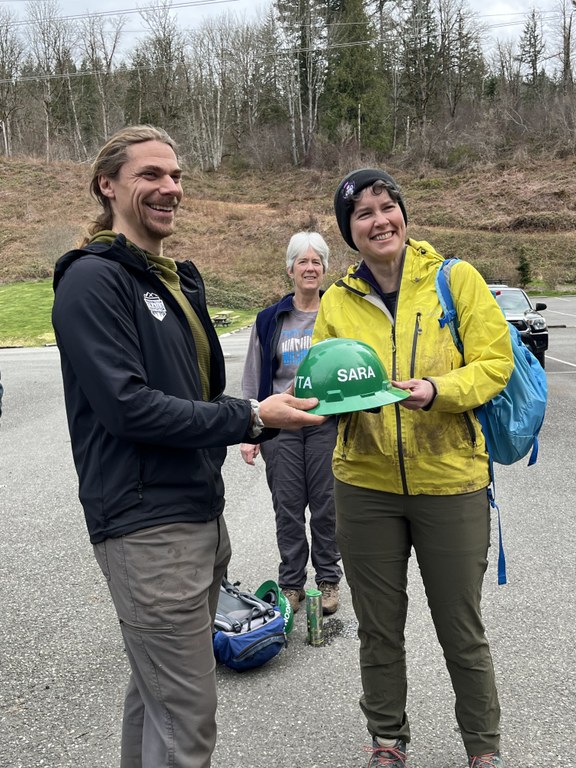 A WTA crew leader and volunteer exchange a green hat in a trailhead parking lot.