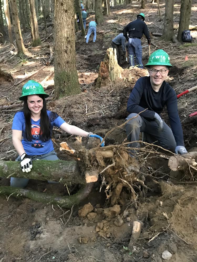 Two volunteers in WTA hard hats remove debris and tree roots from a trail. 