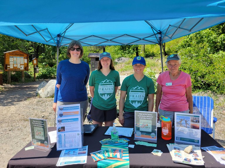 Ingalls Creek Washington Trails Day 2023 Four women, WTA staff and volunteers, pose behind a WTA outreach table at the Ingalls Creek Trailhead