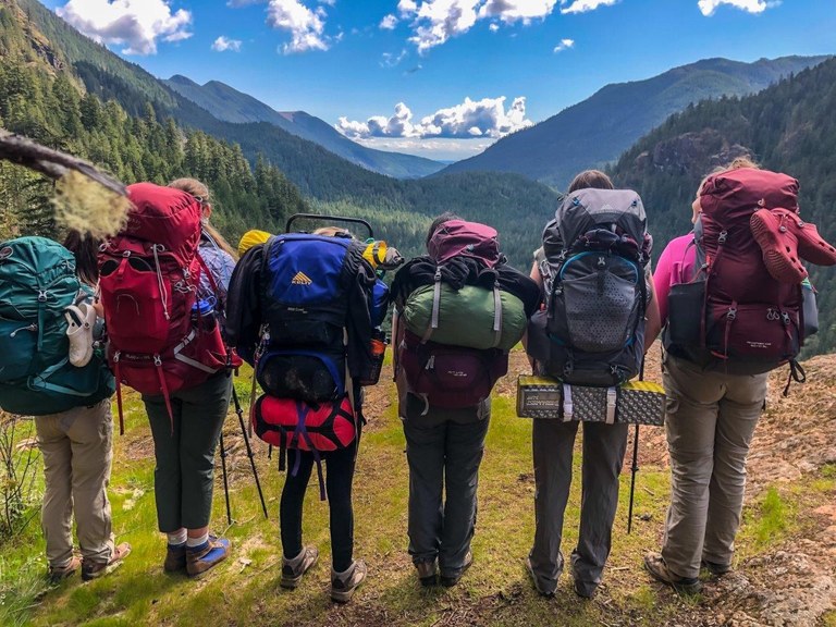A group of youth stand facing away from the camera while wearing packs for an overnight trip. 