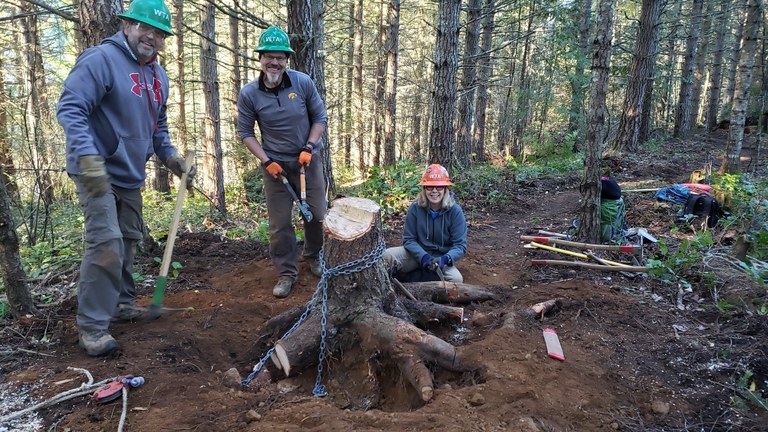 Joni Johnson and two green hats smiling at a chained-up stump ready for removal. Photo by Becca Wanagel Volunteer ACL Joni Johnson and two volunteers smile just before removing a large stump from the Striped Peak Trail.