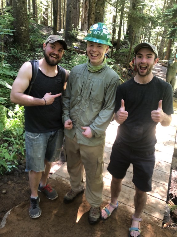 Three people stand on trail, with the person in the middle wearing a hard hat. They are all smiles.