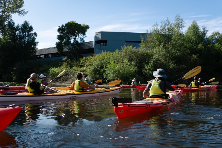 Kayaking A group of people kayaking on a calm river, wearing life vests and hats. Green trees and a modern building are visible in the background under a clear blue sky.