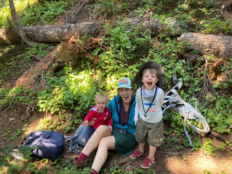A hiker sits on trail with a kid on either side. Everyone is smiling. 
