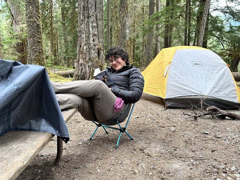 A hiker sits in a camp chair with a book, smiling at the camera. Photo by Jenna van Rijn.