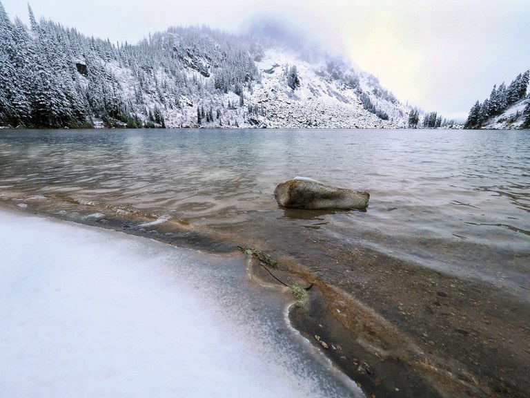 A snowy lakeshore with the frozen surface of a lake in the background. Photo by thebrink