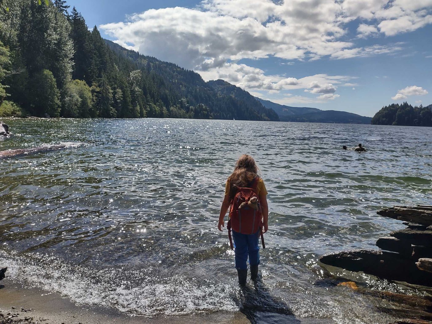 Child wearing a backpack stands in sparkling lake waters on a sunny day