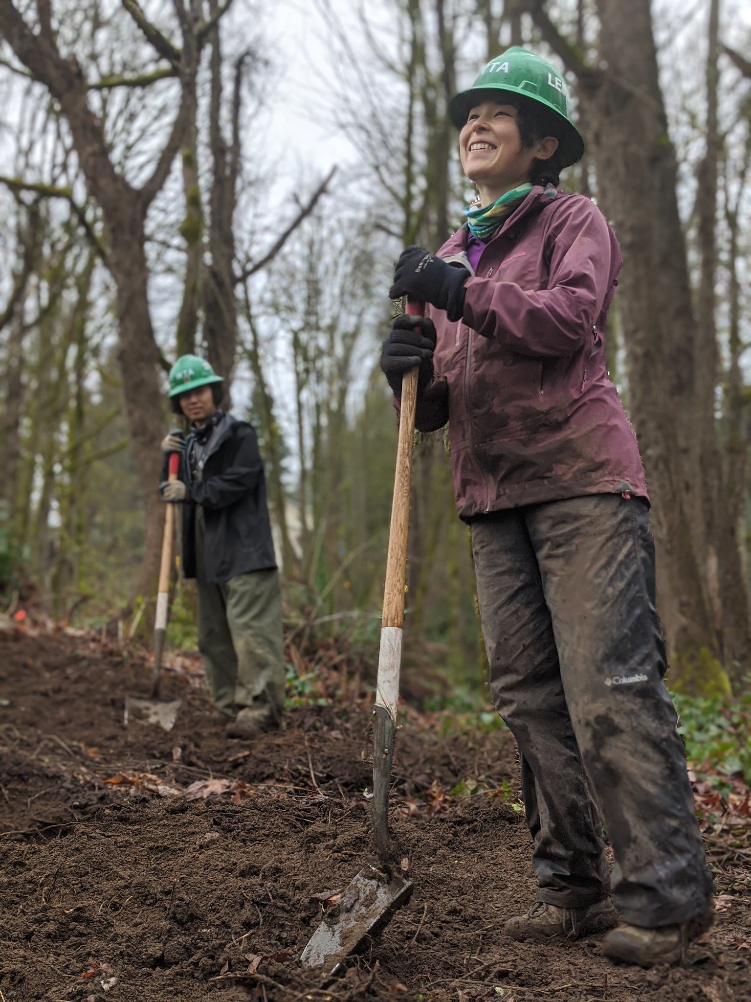A person in muddy rain smiles while holding a shovel and wearing a green WTA hard hat. 