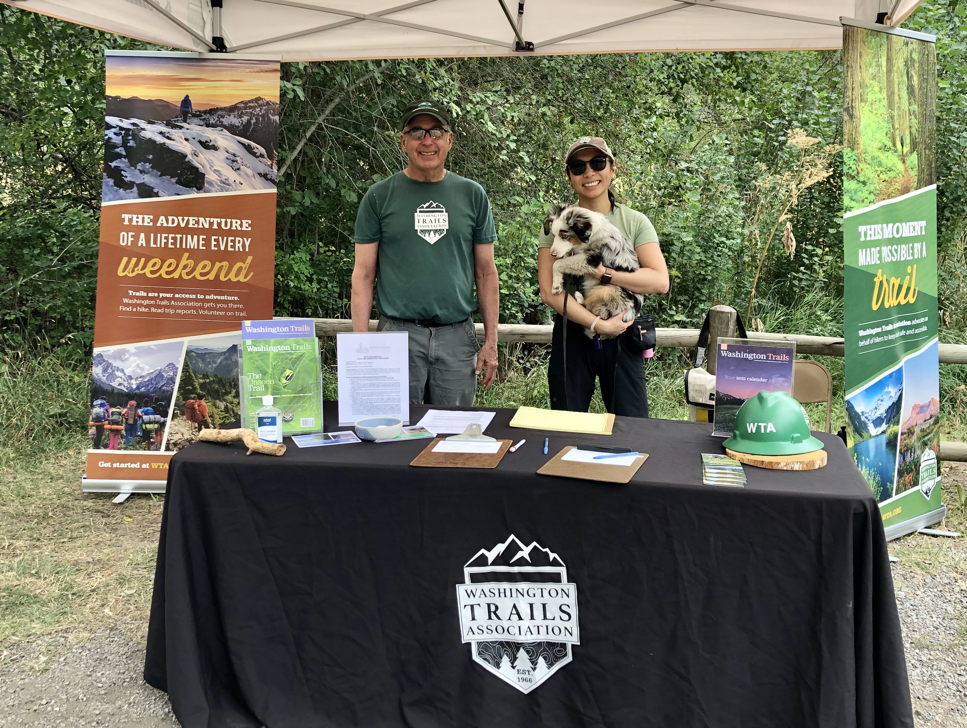 Two volunteers, one holding a puppy, smile from behind a fully stocked WTA table at the Liberty Lake trailhead.