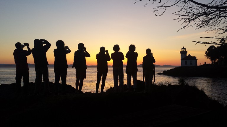 A group of people stand silhouetted facing a sunset, many taking photos of the colorful water beyond.