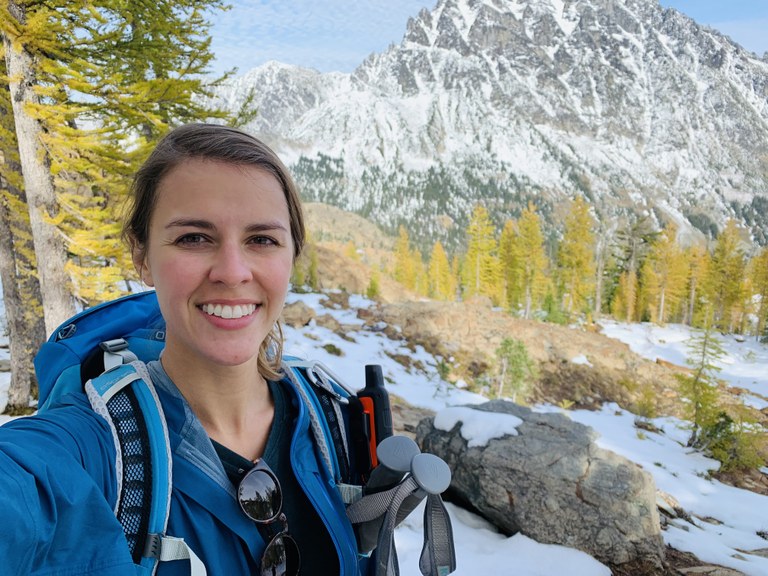 WTA member Lindsay M Lindsay smiles at the camera while wearing a backpack and standing on trail in front of golden larches and a rocky peak.