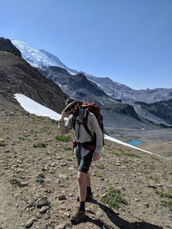 WTA member Logan R Logan stands on a rocky pass and smiles at the camera. A faint trail can be seen in the valley below him, and a sliver of Mount Rainier is peeking out on the left.