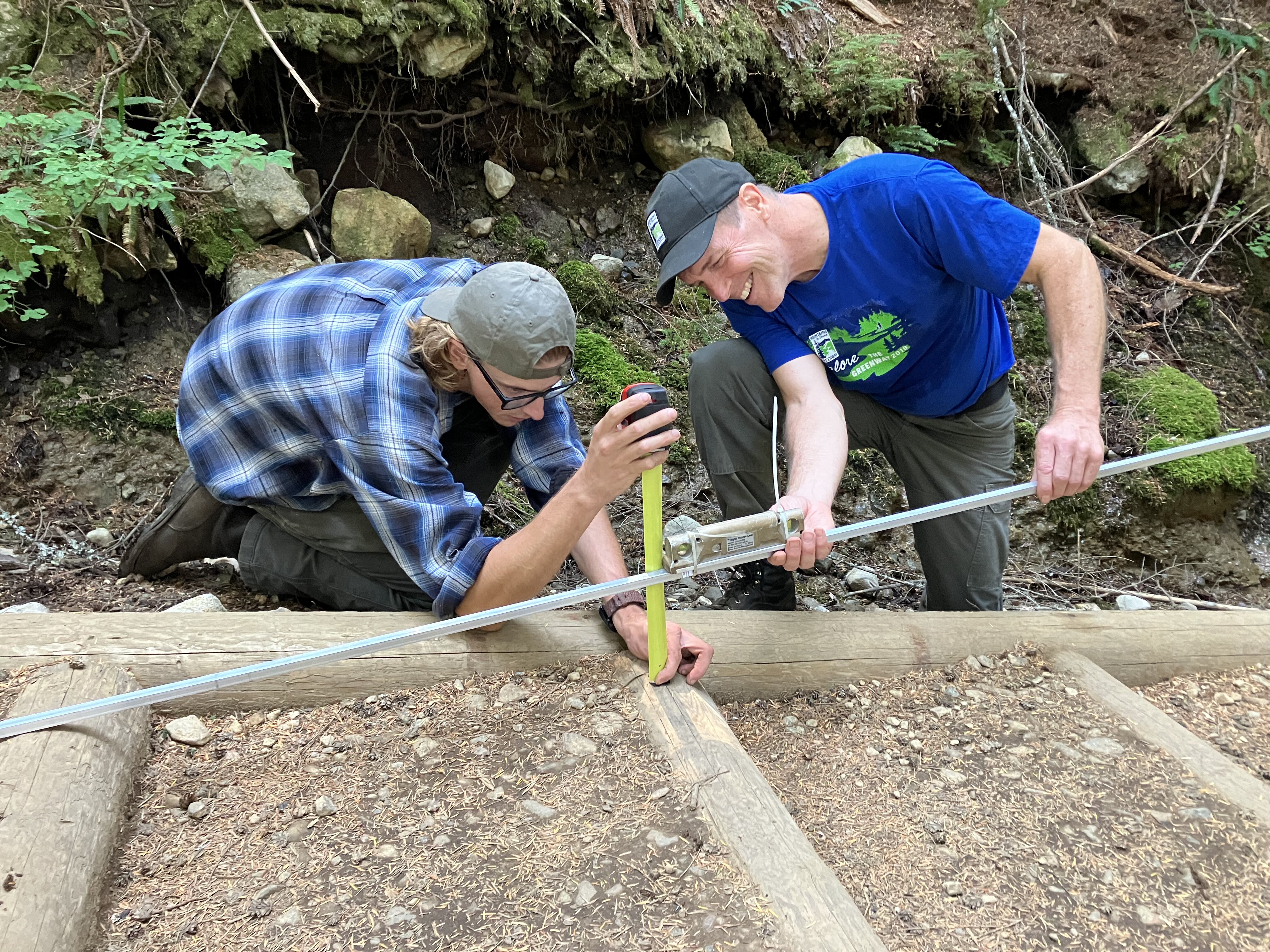 Two people, one of them smiling, kneel near a trail to measure it with tools