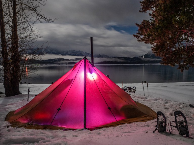 Photo by Michael Margoshes. A tent is pitched on the shore of a snowy lake. Outside the tent are snowshoes and trekking poles. Clouds drift over the lake in the background.