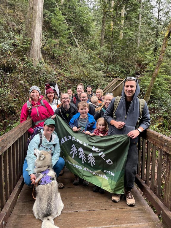 Group of 14 people stand on a bridge in the woods, holding a military wild banner, a dog sits in the front