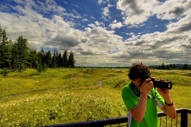 Mima Mounds. Devin Sawyer. A person taking a photo at the end of a boardwalk with a green field behind them.