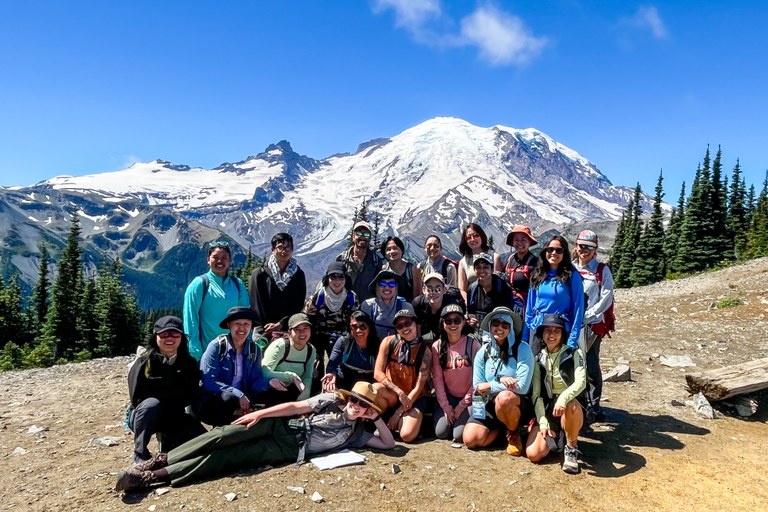 Mount Rainier group photo A group of people poses happily outdoors in front of a snow-capped mountain, with clear blue skies and pine trees in the background, creating a joyful and adventurous atmosphere.