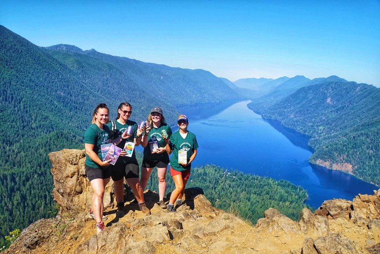Four hikers wearing matching green shirts and holding books and drinks pose for a photo at the top of a rocky outcropping. A lake and evergreen-covered hills are visible far below.