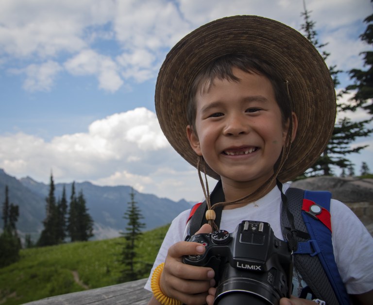 A boy with a camera and sun hat smiles.