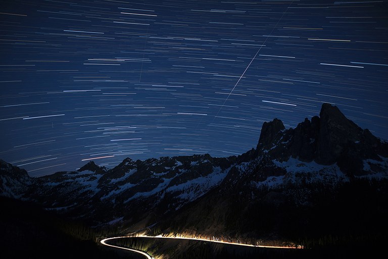 A starry sky and mountains with an illuminated highway curving around the base of the mountains. 