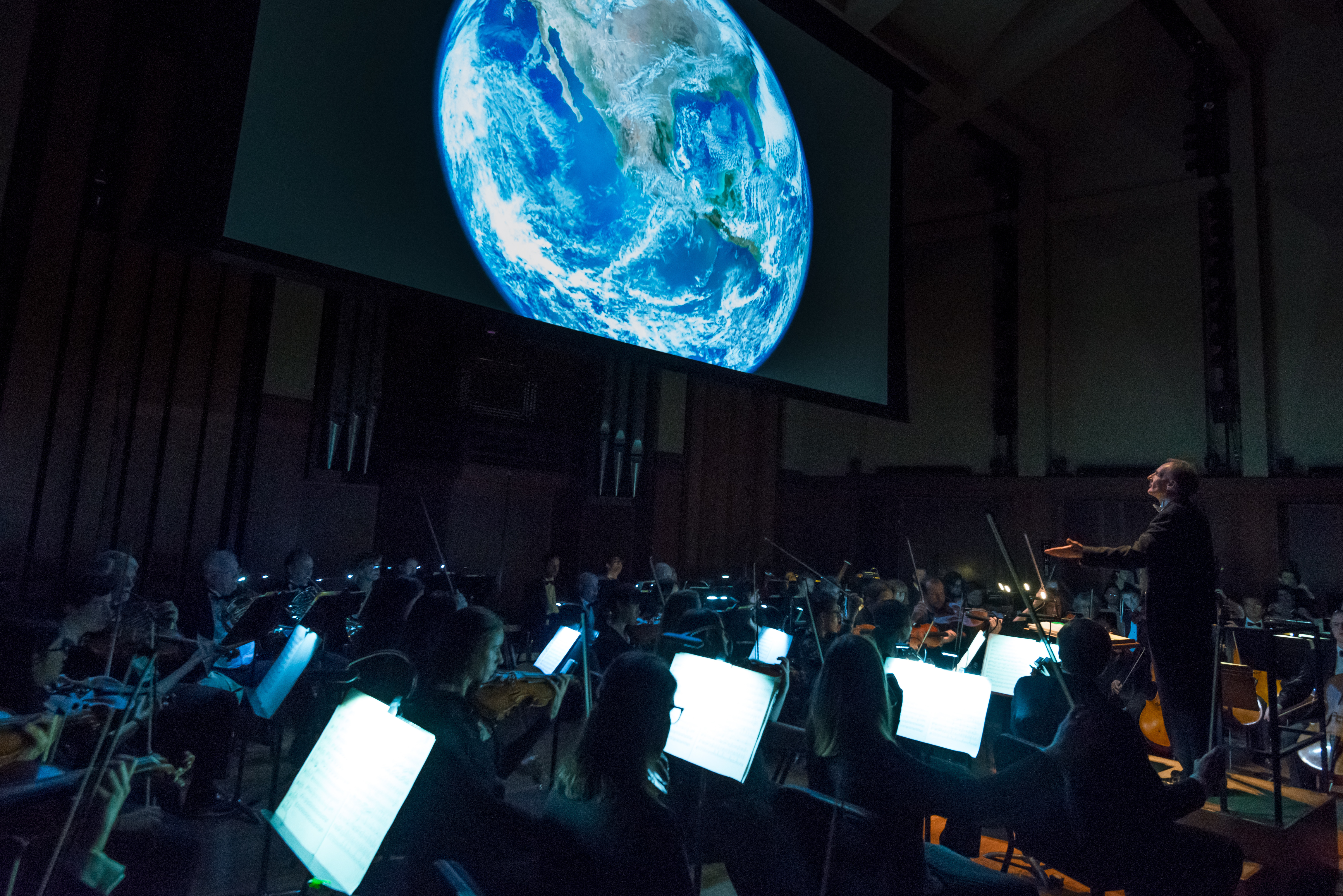 Symphony preforms in front of screen with picture of earth from space.