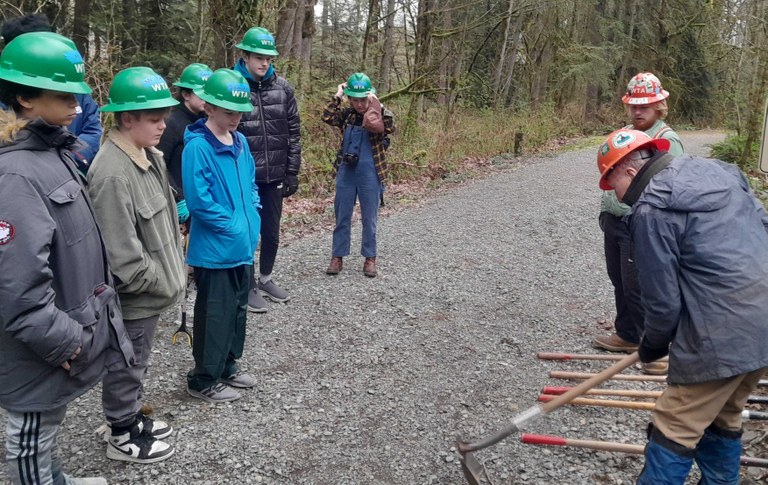 A group of kids on the left of the image watch two people demonstrate how to use various tools. 