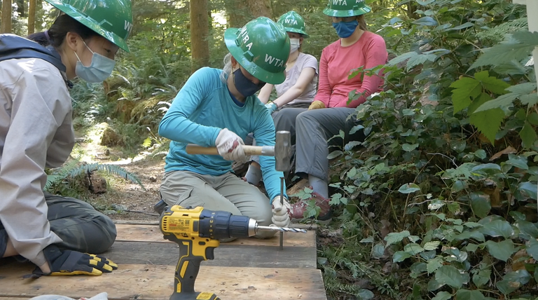 Youth volunteer vacation by Cole Hanych. A teen volunteer uses a hammer while other volunteers watch. Everyone is wearing green hard hats and face coverings.