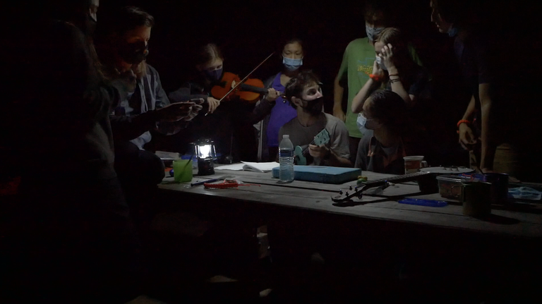 youth volunteer vacation. Photo by Sarah Williams. Teens stand around a picnic table in the dark, lit by a lantern. Teens are playing stringed instruments, and a few more instruments sit on the table. Everyone is wearing face coverings.