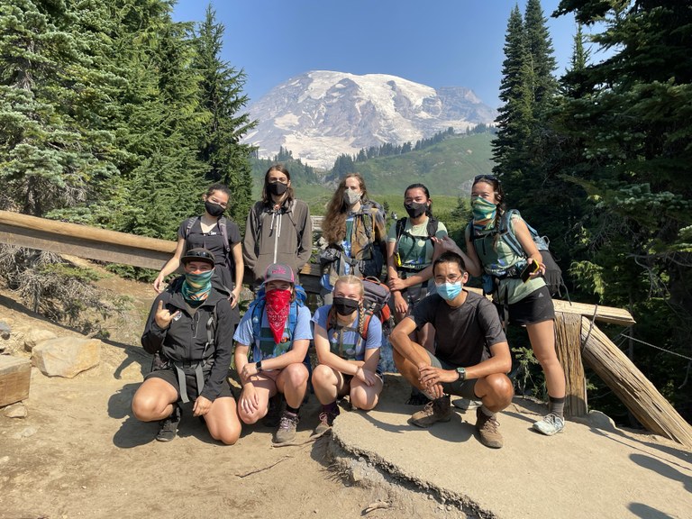 Youth volunteer vacation by WTA staff. A group of nine teens, all wearing face coverings, pose for a photo in front of a wooden fence with Mount Rainier in the background on a sunny day.